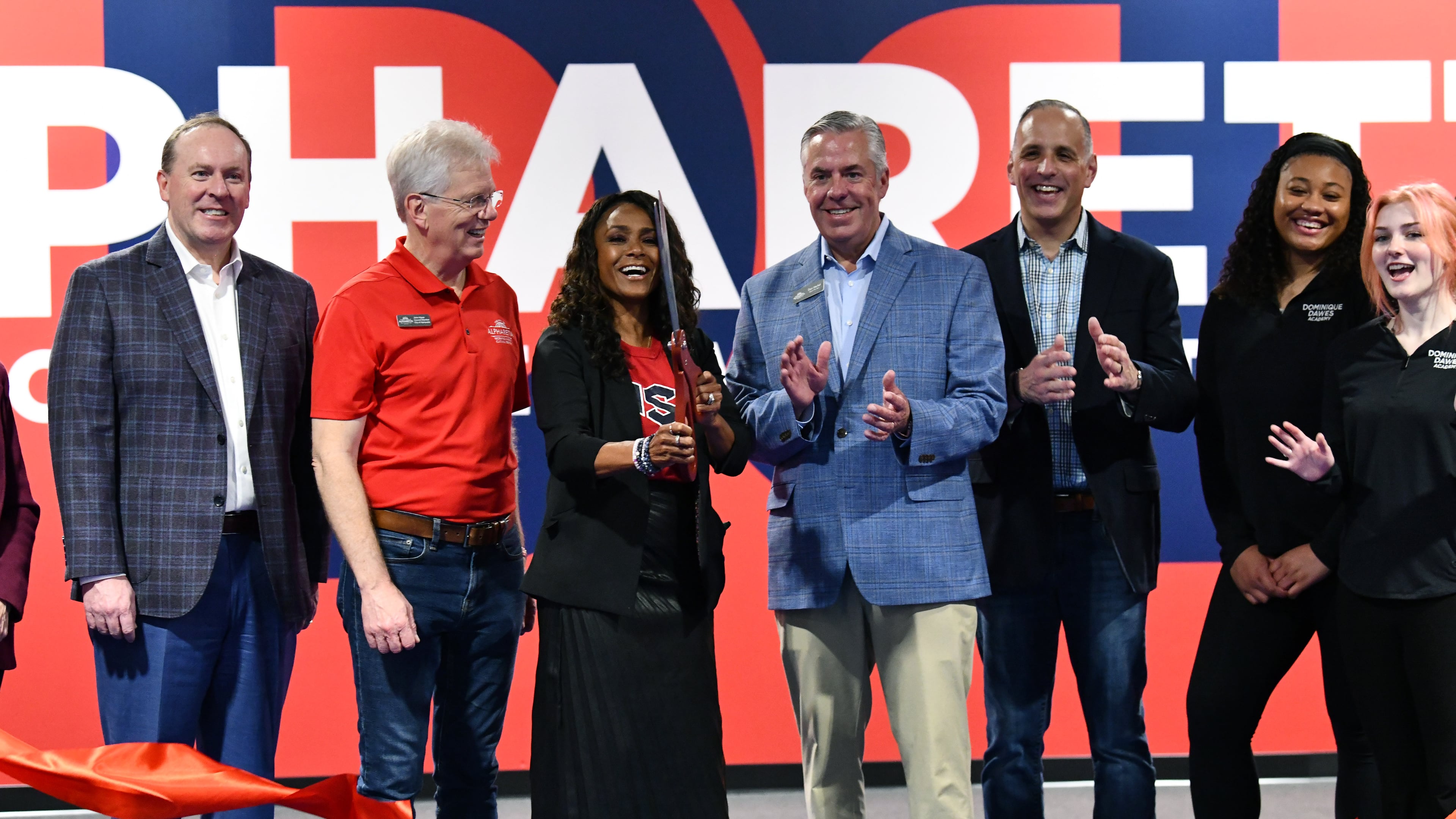 Dominique Dawes holding ribbon-cutting scissors reacts as she stands with VIP guests during the opening day of Dominique Dawes Academy, Friday, March 6, 2026, in Alpharetta. Dawes, a 1996 Olympics medal winning gymnast, has seven gymnastics academies in Maryland and Texas and now has expanded to metro Atlanta, in Alpharetta. (Hyosub Shin/AJC)