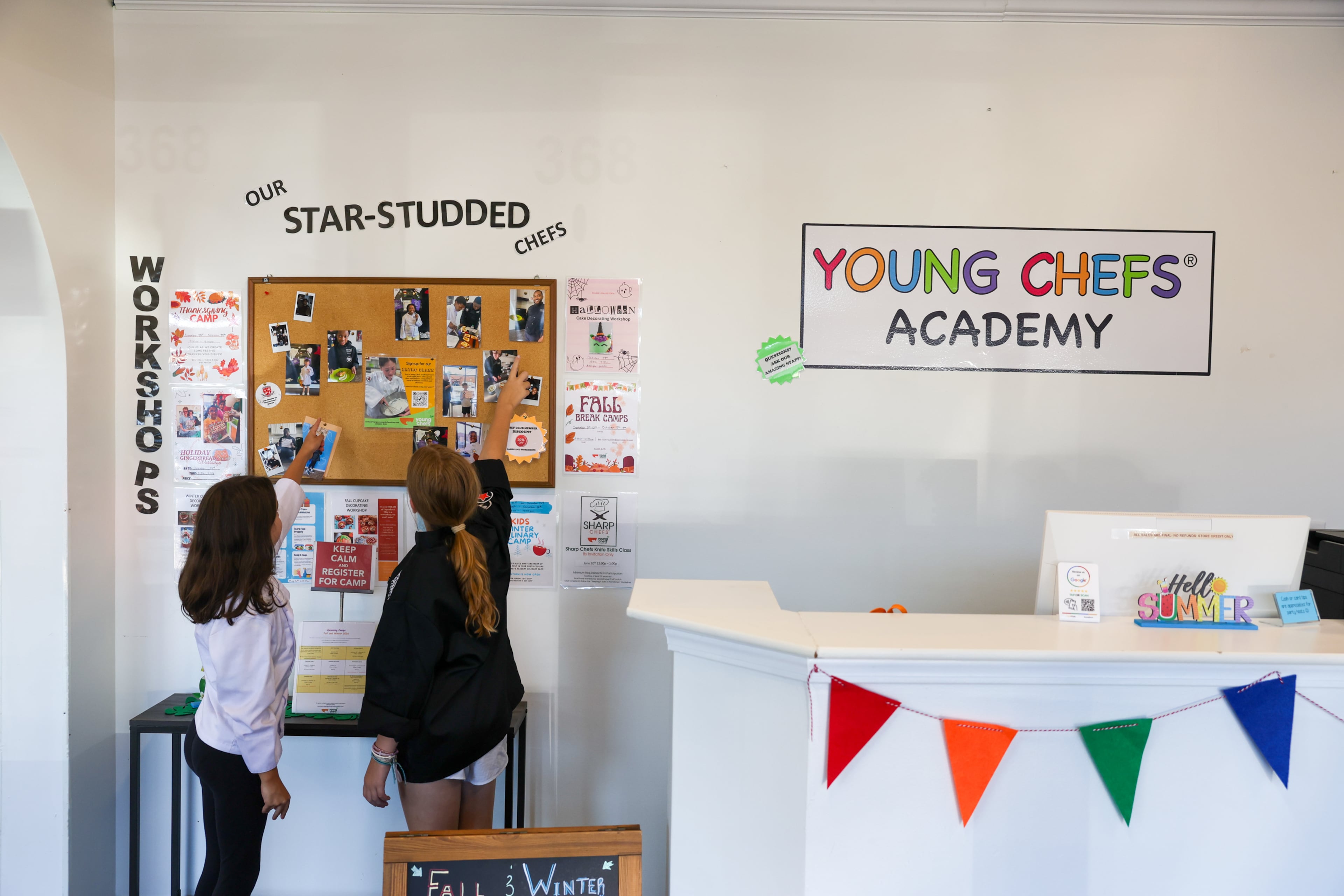 (L-R) Students Violet (last name not provided), 10, and Colette Casey, 9, point to a board of pictures at Young Chefs Academy in Sandy Springs on Aug. 20, 2026. (Arvin Temkar/AJC)